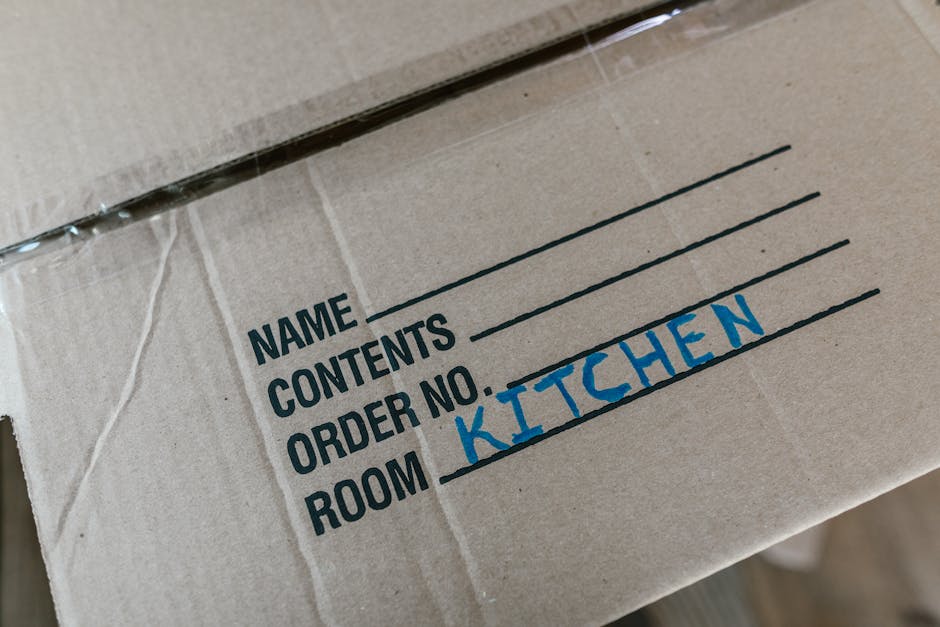Close-up of a cardboard moving box with printed labels indicating sections for 'Name', 'Contents', 'Order No', and 'Room'. The handwritten label 'Kitchen' is visible in blue ink under the 'Room' secti